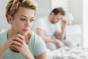 In the foreground, a woman sits in concerned thought. In the background, her partner sits with his head in his hand. They are frustrated about vaginal dryness during menopause and its effects on their intimacy.